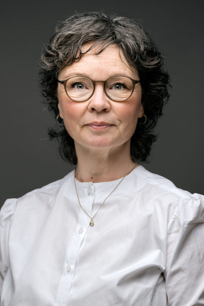 En kvinde med kort, krøllet hår og briller står mod en mørk baggrund, iført en hvid bluse og en delikat halskæde. Hun fremstår fokuseret og fattet.