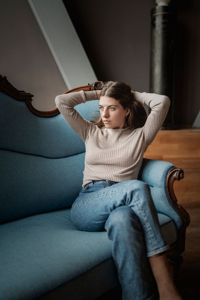 En kvinde i beige sweater og blå jeans sidder i en blå vintagesofa med armene hvilende bag hovedet og kigger eftertænksomt til siden. Baggrunden består af en trætrappe og en mørk søjle.