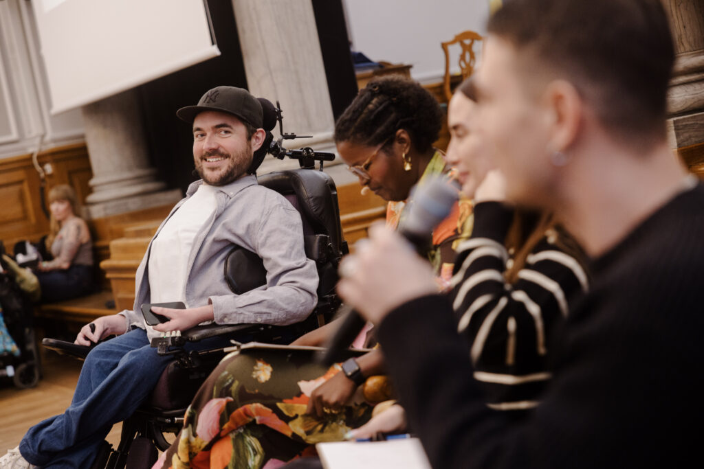 En gruppe på fire personer er engageret i en paneldebat. En person, der smiler, sidder i kørestol. De er indendørs, siddende foran en væg med træpaneler. Da samtalen bliver til "Min pause fra reels – og hvorfor du også skulle prøve det," holder en anden en mikrofon, mens andre lytter opmærksomt.