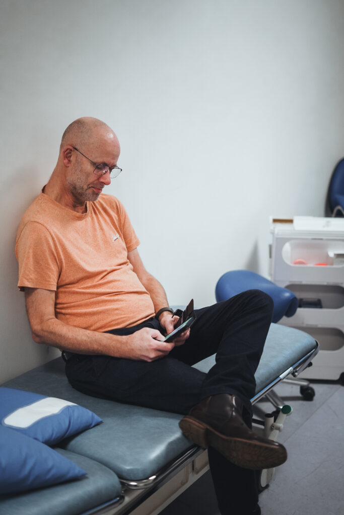 En midaldrende mand iført briller og en orange t-shirt sidder på en lægeundersøgelsesseng og kigger ned på sin telefon i et klinikrum. En blå stol og medicinsk udstyr er i baggrunden.