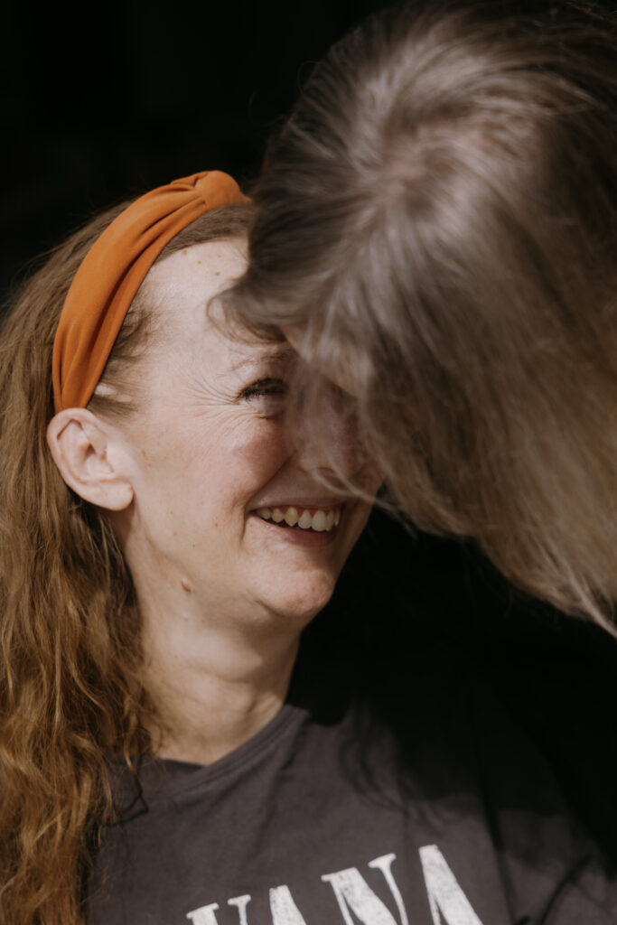 En kvinde med langt brunt hår og et orange pandebånd smiler varmt, mens en anden person med gråt hår læner sig tættere på og delvist skjuler hendes ansigt. Stemningen er intim og glædelig.