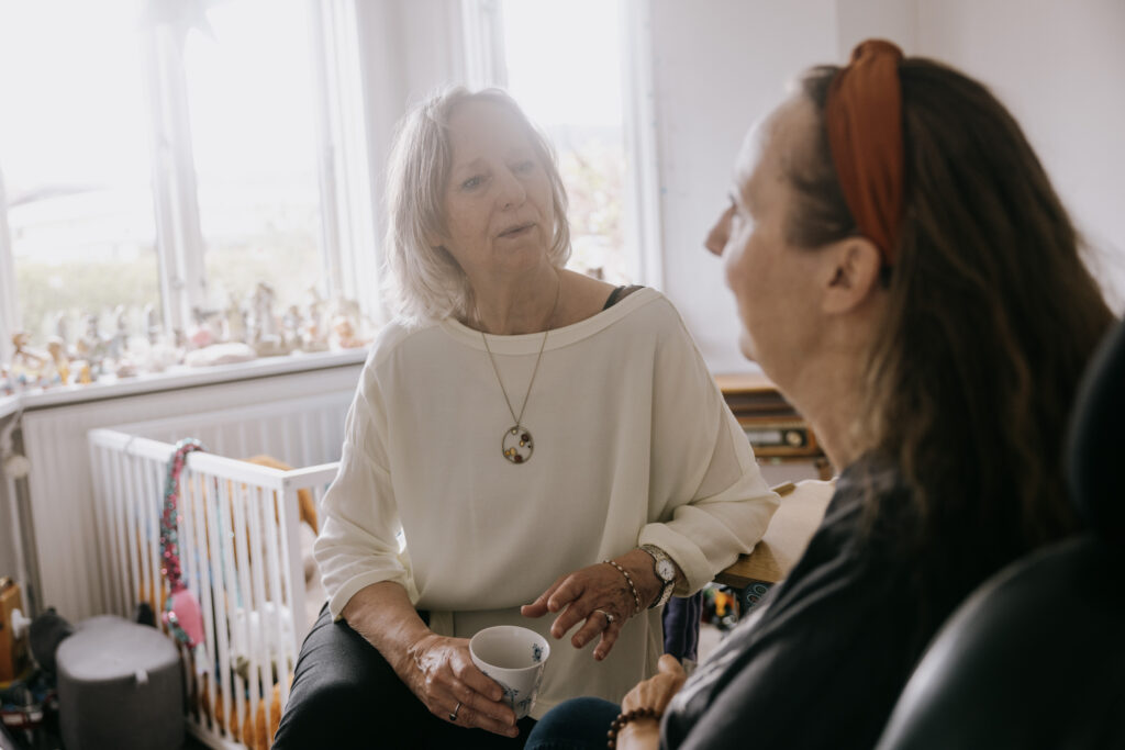 To kvinder sidder indendørs og taler. Den ene kvinde med gråt hår holder en kop og gestikulerer, mens hun taler, mens den anden lytter. Naturligt lys kommer ind fra et vindue bag dem, og forskellige små ornamenter hænger i vindueskarmen.