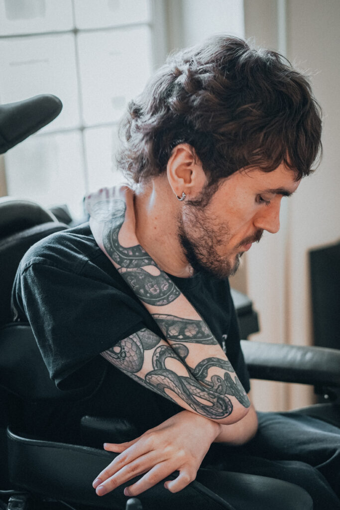 En mand med skæg og kort hår, iført en sort t-shirt, sidder i en kørestol. Han har en detaljeret tatovering på armen, som hviler hen over hans bryst. Baggrunden viser et vindue med naturligt lys.
