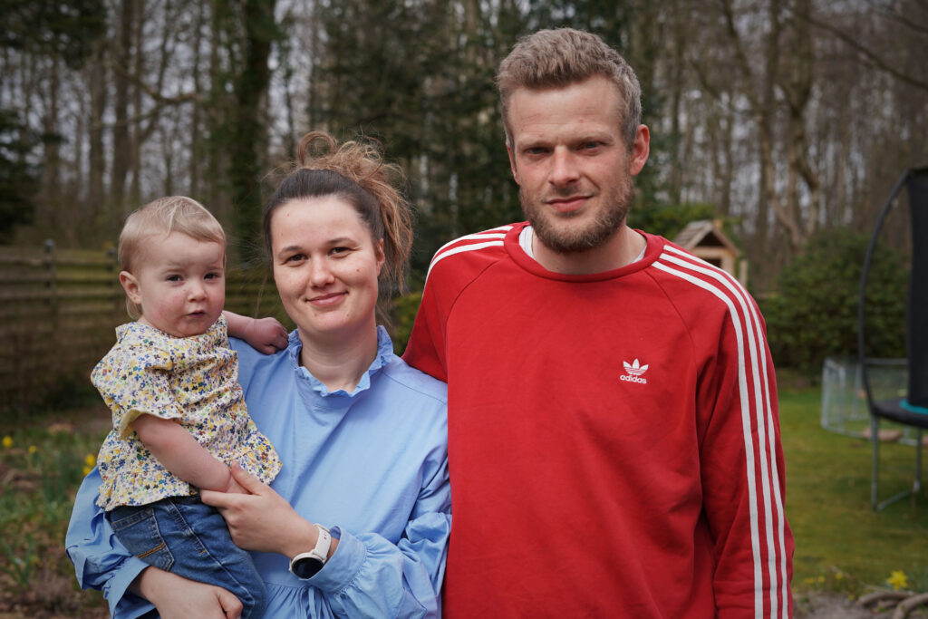 En kvinde, der holder en baby, står ved siden af en mand i en rød Adidas-sweatshirt. De er udendørs i en have med træer og et træhegn i baggrunden. Alle tre ser mod kameraet.