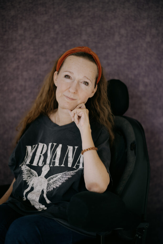 En kvinde med langt brunt hår, iført en Nirvana t-shirt og et rødt pandebånd, sidder i en kørestol mod en lilla tekstureret baggrund, hviler hagen på hånden og smiler blidt til kameraet.