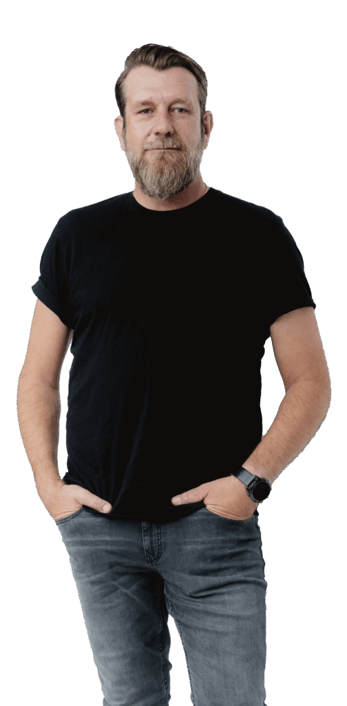 En mand med kort hår og skæg står med ansigtet fremad, iført en sort t-shirt, grå jeans og et smartwatch. Han har hænderne i lommerne og smiler let.