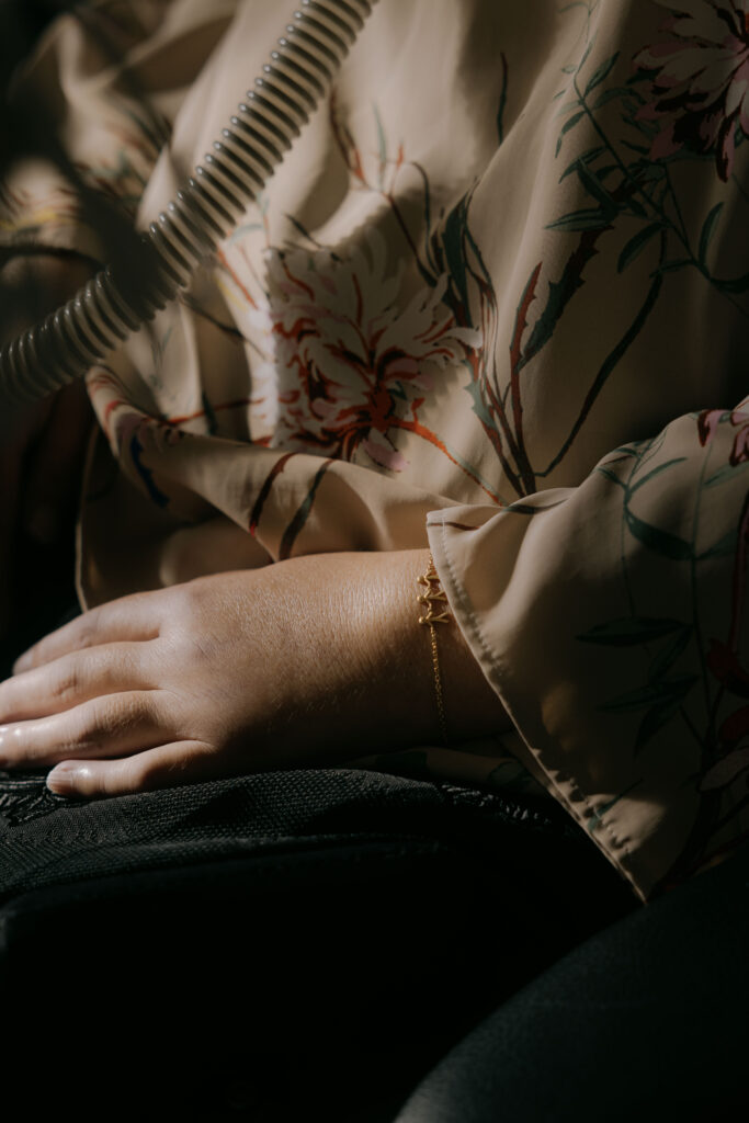 Et nærbillede af en persons hånd, der hviler på skødet, iført et guldarmbånd. Personen er klædt i en blomstermønstret, beige bluse, og en del af et snoet rør er synlig i baggrunden.