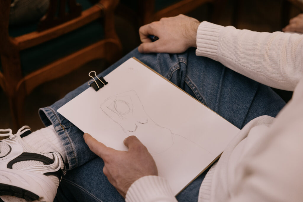 En person iført jeans og en hvid sweater tegner skitser på et udklipsholder, der hviler på deres skød. Tegningen ser ud til at være en omrids af en flaske eller lignende genstand. Personen holder en blyant og har hvide sneakers på.