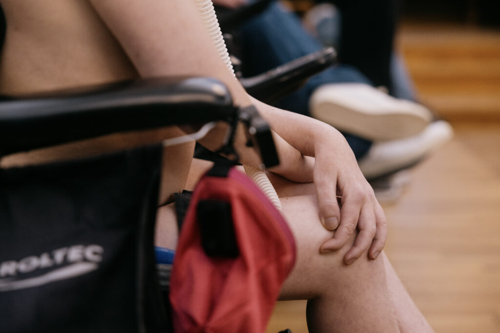 En person sidder i en kørestol med armen hvilende på knæet. En anden person i baggrunden er lidt ude af fokus og sidder med benene over kors.