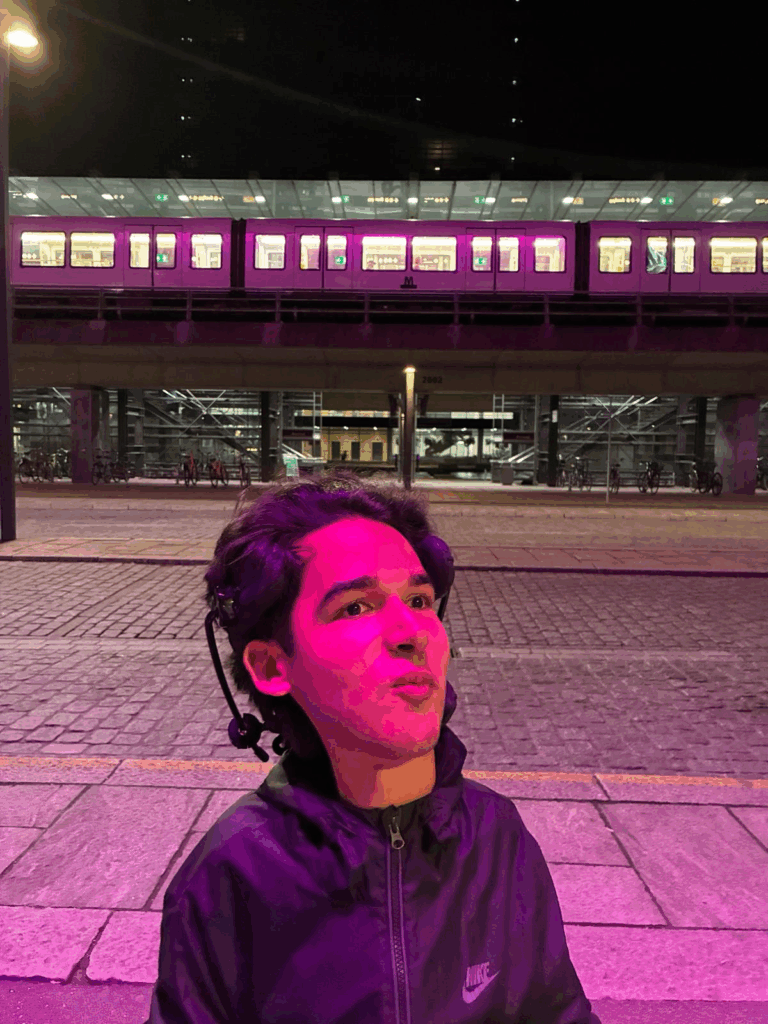 En ung mand med mørkt hår og headset kigger opad, oplyst af lyserødt-lilla lys om natten, med et moderne, oplyst tog og perron i baggrunden.
