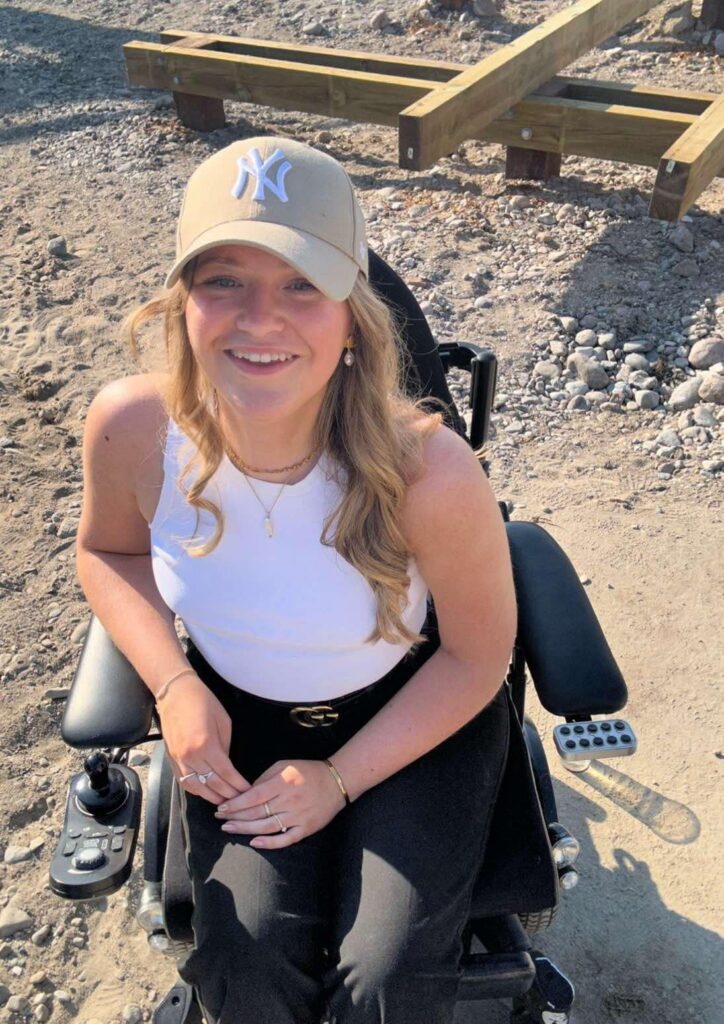 En ung kvinde med langt blond hår, iført en brun Yankees-kasket og en hvid ærmeløs top, smiler, mens hun sidder i en motoriseret kørestol udendørs på en solskinsdag. Træbjælker og grus er synlige i baggrunden.