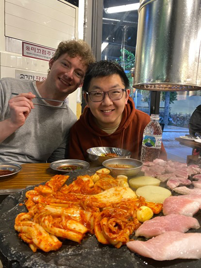 To personer smiler ved et restaurantbord med en koreansk BBQ foran sig, hvor de tilbereder grøntsager, kimchi og kødskiver. Der hænger en ventil over grillen, og der er vand og krydderier på bordet.