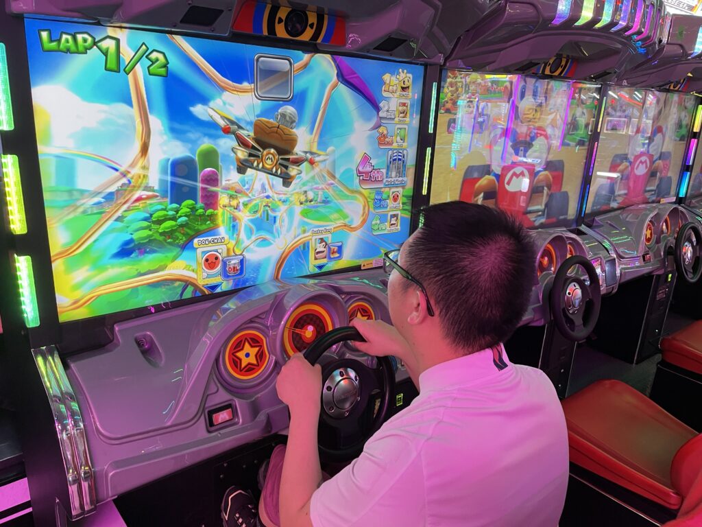 En person med briller og hvid skjorte spiller et Mario Kart arkadespil og holder fast i rattet, mens farverig spilgrafik vises på skærmen foran dem.