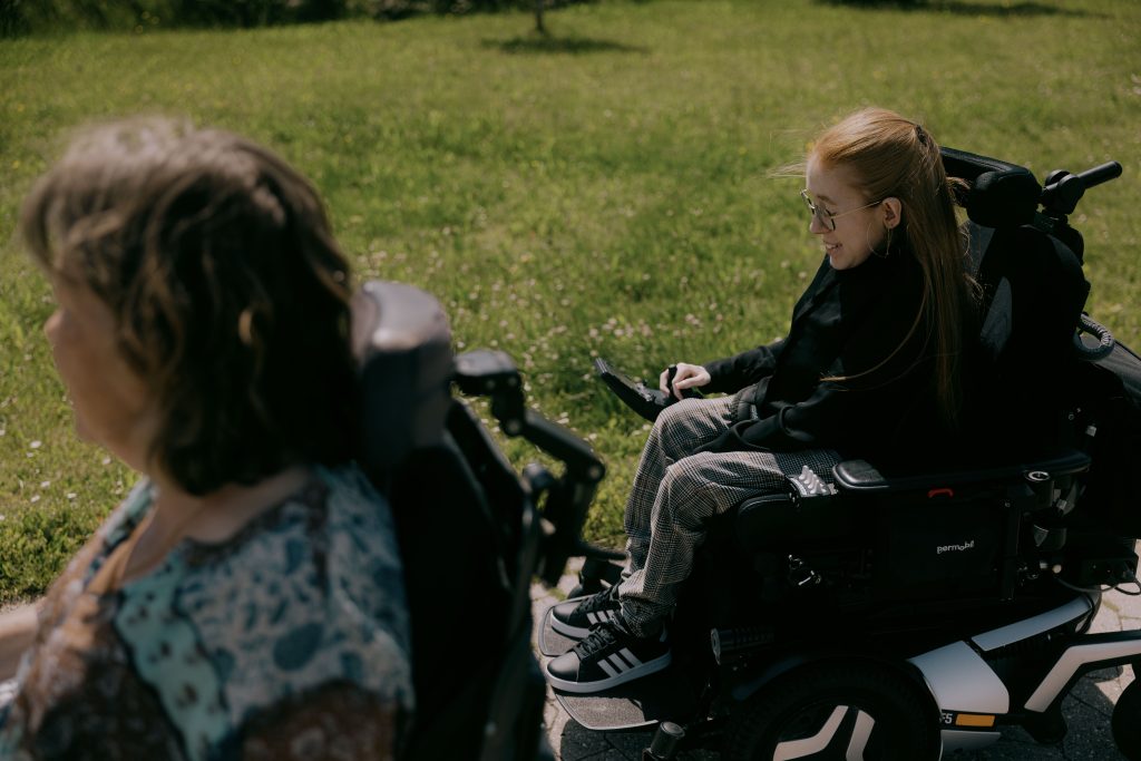 To kvinder i motoriserede kørestole er udendørs på en solskinsdag. Den ene er i forgrunden, delvist ude af fokus, og den anden smiler i baggrunden, omgivet af grønt græs.