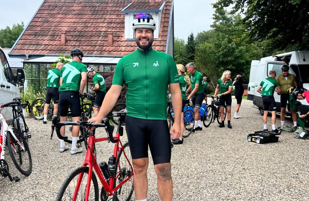En smilende mand i cykeltøj står med en rød cykel foran en gruppe cyklister, alle iført matchende grønne trøjer, uden for en bygning med rødt tegltag. Andre gør cykler klar og taler i baggrunden.
