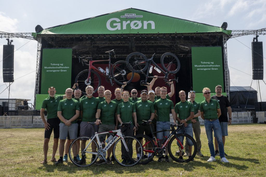 En gruppe mennesker i matchende grønne skjorter står foran en scene med et grønt tag med påskriften "Grøn". Flere cykler står foran, og nogle holdes over deres hoveder. Det ser ud til at være en udendørs begivenhed eller koncert.