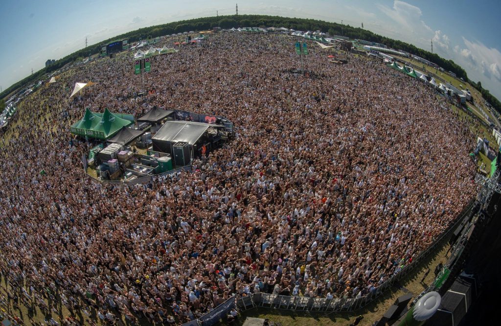 Et fiskeøje-billede af en stor udendørs musikfestival, hvor publikum er samlet foran en stor scene, omgivet af telte og grønne områder under en klar, solrig himmel.