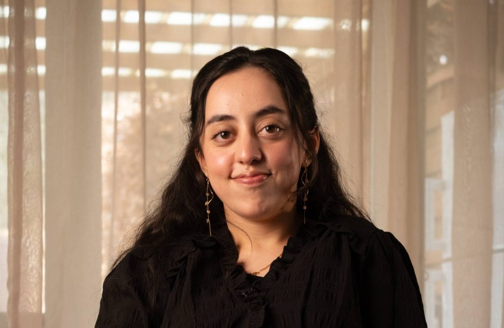 En person med langt mørkt hår, iført sort top og guldøreringe, smiler blidt, mens hun står indendørs foran gennemsigtige gardiner, hvor sollyset trænger igennem.