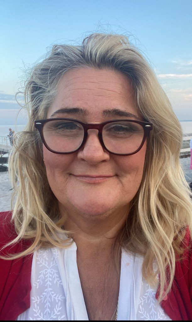 En kvinde med blondt hår og briller smiler til kameraet på en strand ved solnedgang iført en rød sweater over en hvid top. Himlen og vandet er synligt i baggrunden.
