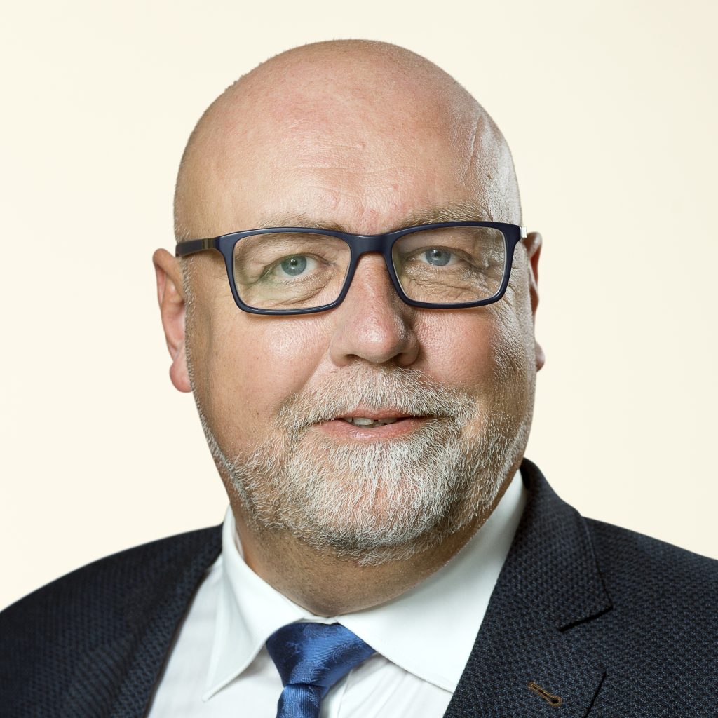 En midaldrende skaldet mand med briller, kort gråt skæg og overskæg, iført mørkt jakkesæt, hvid skjorte og blåt slips, smiler let mod en almindelig lys baggrund.