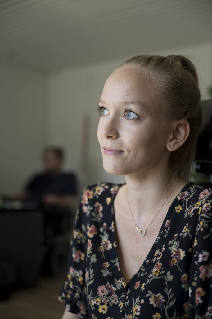 En kvinde med blondt hår i hestehale og en blomstret top kigger eftertænksomt til siden i et svagt oplyst rum; en mand sidder i baggrunden, lidt ude af fokus.