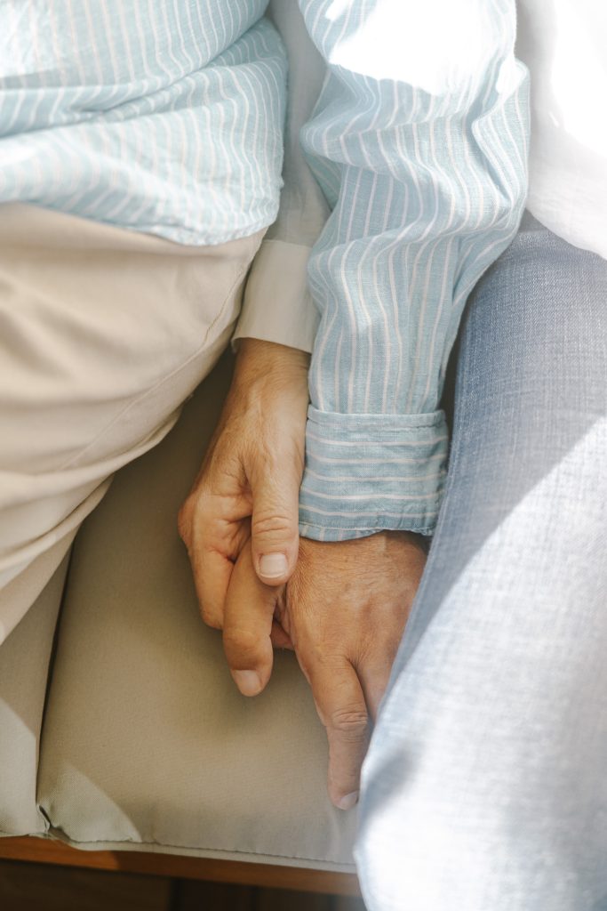 To mennesker, der sidder tæt sammen, holder forsigtigt hinanden i hånden. Den ene har lyse beige bukser og en blåstribet skjorte på, den anden har lyseblå jeans på. Sollyset belyser blidt deres hænder og arme.