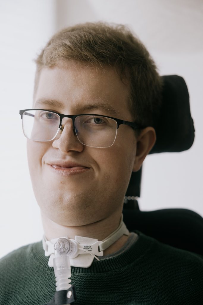 En ung mand med kort hår, briller og en grøn sweater bruger en respirator med en trakeostomislange. Han sidder i en kørestol, kigger ind i kameraet og smiler lidt.