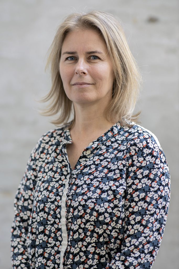 En kvinde med skulderlangt blondt hår bærer en blomstermønstret skjorte med knapper og står foran en lys, sløret baggrund og ser roligt ind i kameraet.
