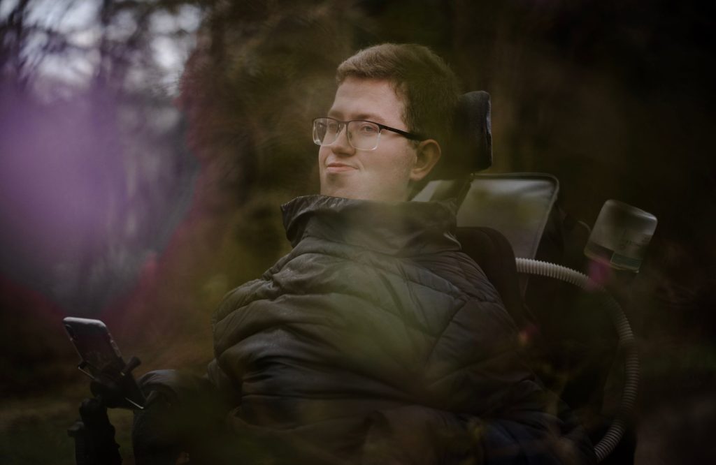 En person med briller og en mørk jakke sidder i en kørestol udendørs, omgivet af planter med blødt fokus og naturligt lys. Personen kigger ud til siden med et lille smil, og et hjælpemiddel er synligt.