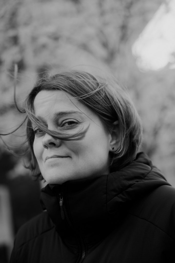 En kvinde med kort hår iført en mørk jakke står udendørs. Hårstråene blæser hen over hendes ansigt, og hun har et lille smil på læben. Baggrunden er sløret, og billedet er i sort-hvid.
