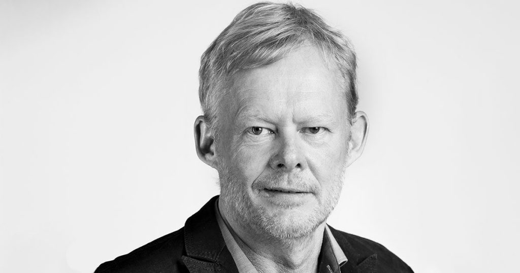 En midaldrende mand med lyst hår og skæg iført mørk blazer og lys skjorte ser direkte ind i kameraet med et neutralt udtryk mod en ensfarvet baggrund.