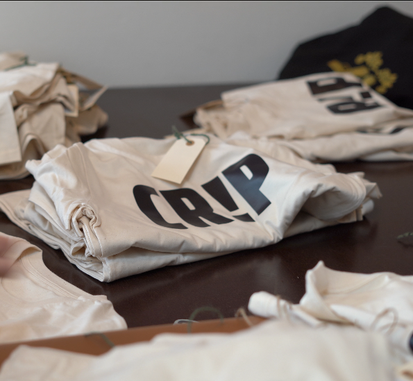 En stak foldede hvide t-shirts med ordet "CRIP" i fede sorte bogstaver ligger på et bord, omgivet af andre BPA-fri t-shirts og tags, der er synlige i baggrunden.