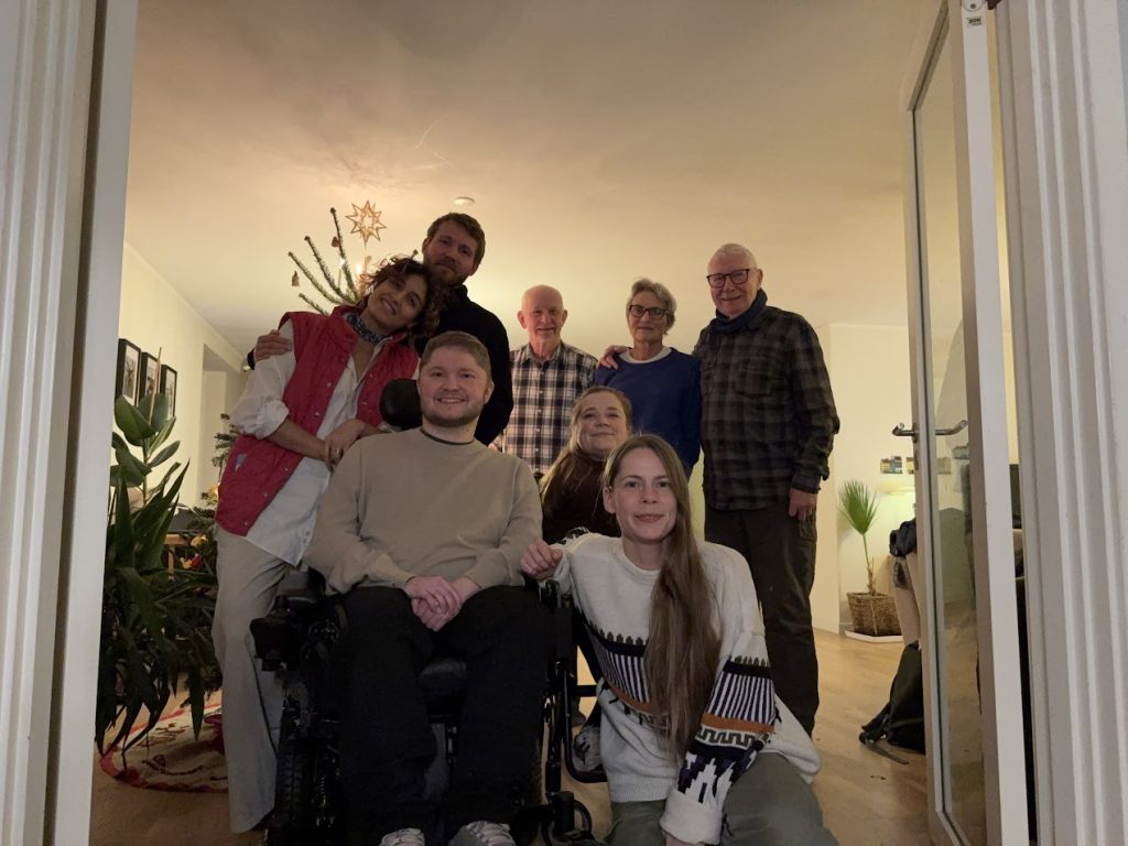 Otte voksne, heriblandt en mand i kørestol, poserer og smiler sammen i en varmt oplyst stue med stueplanter og et pyntet juletræ i baggrunden.