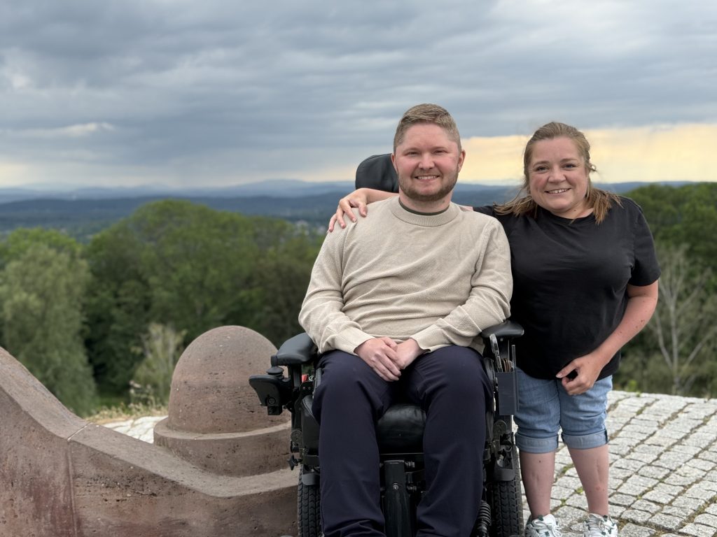 En smilende mand i kørestol og en kvinde står tæt sammen udendørs, med hendes arm om hans skulder. De står på en udkigspost med træer og bakker i baggrunden under en overskyet himmel.