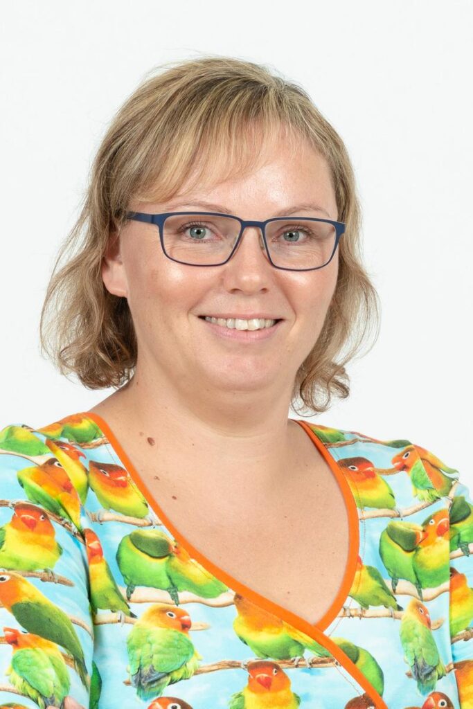 En kvinde med kort blondt hår og briller smiler til kameraet. Hun er iført en farverig skjorte med et mønster af grønne, orange og gule papegøjer og en orange kant. Baggrunden er helt hvid.
