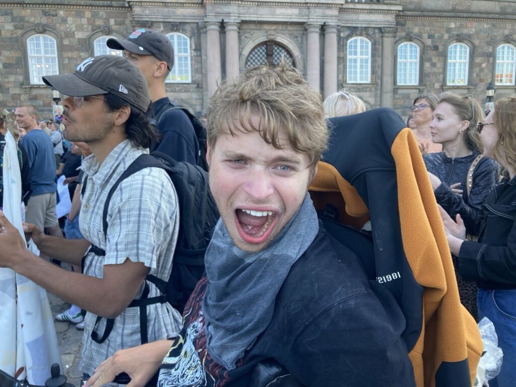 En ung mand med lysebrunt hår i kørestol smiler bredt ved en udendørs sammenkomst. Folk står omkring ham, nogle klapper, og i baggrunden ses en historisk bygning under en klar himmel.