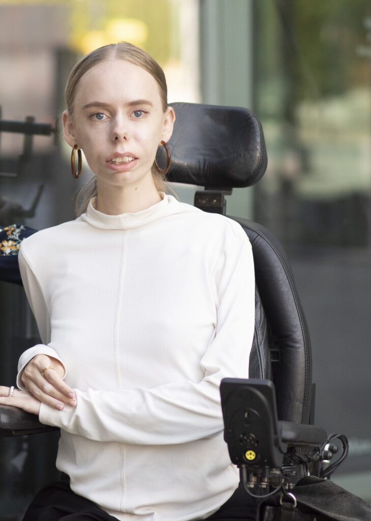 En ung kvinde med lys hud og blondt hår, iført en hvid langærmet skjorte og store øreringe, sidder i en el-kørestol udendørs og kigger selvsikkert ind i kameraet.