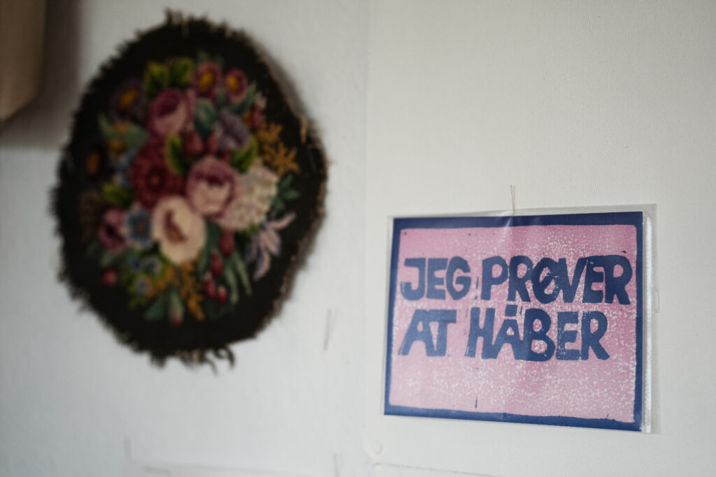 Et indrammet skilt med de danske ord "JEG PRØVER AT HÅBER" med store blå bogstaver hænger på væggen ved siden af et rundt, blomstret broderikunstværk.