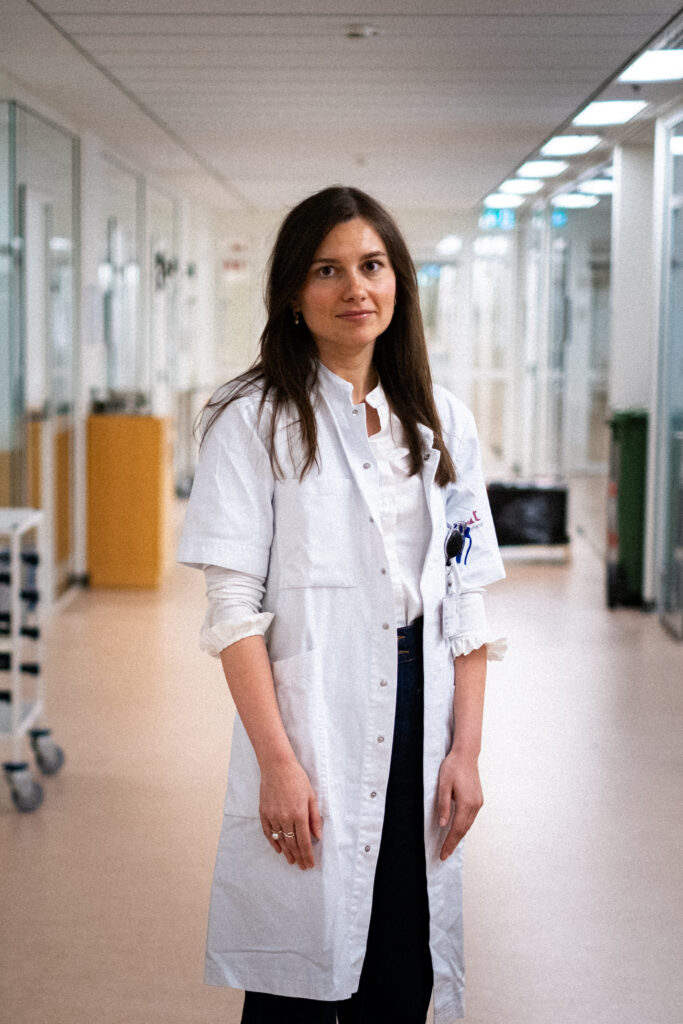 En kvinde iført en hvid kittel står i en lys gang med glasvægge og ser ind i kameraet med et neutralt udtryk. Omgivelserne ligner et hospital eller en medicinsk institution.
