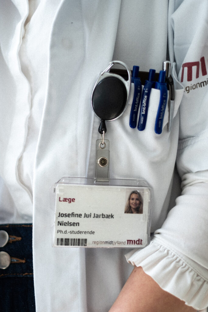 Et nærbillede af en person iført en hvid kittel med kuglepenne i lommen og et ID-skilt med navnet Josefine Jui Jarbæk Nielsen, en ph.d.-studerende læge, fastgjort til kittelen.