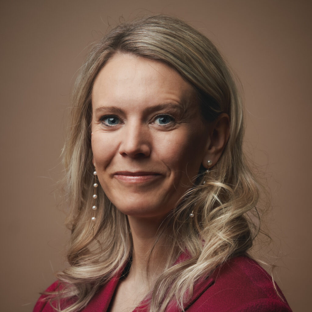 En kvinde med langt, blondt, bølget hår, iført en rød blazer og perleøreringe, smiler blidt foran en ensfarvet brun baggrund.