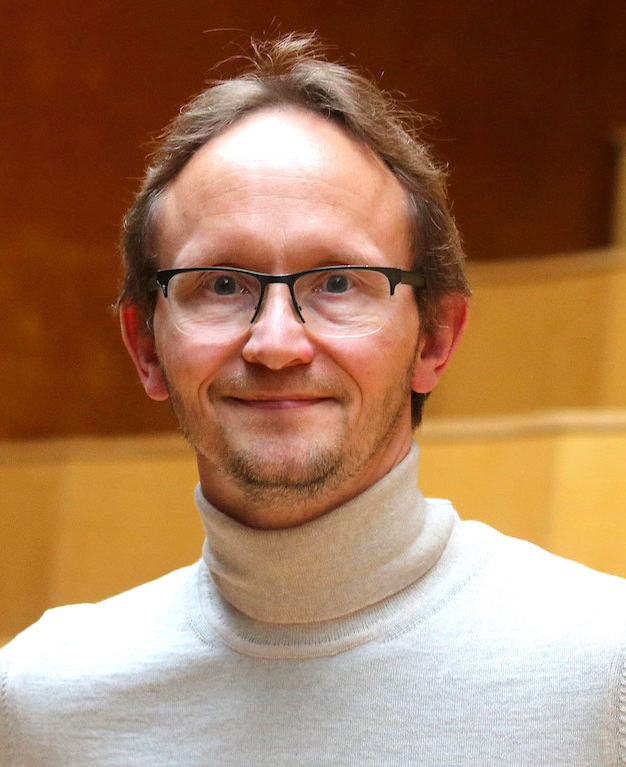 En mand med kort, lysebrunt hår og briller smiler til kameraet. Han har en lysegrå rullekravesweater på og står indendørs foran en baggrund i varme farver, der er lidt sløret.