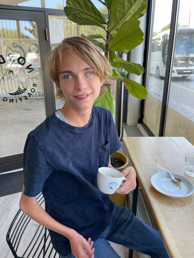 En ung person med lysebrunt hår sidder ved et cafébord, holder en hvid kop i hånden og smiler. Der står en tom tallerken og et glas vand på bordet, med en stor grøn plante og vinduer i baggrunden.