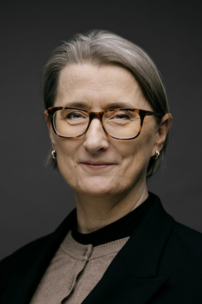 En person i middelalderen med kort, gråt hår, iført skildpaddeskalsbriller, små ringøreringe, en sort blazer og en beige bluse, står med et neutralt udtryk foran en mørk baggrund.