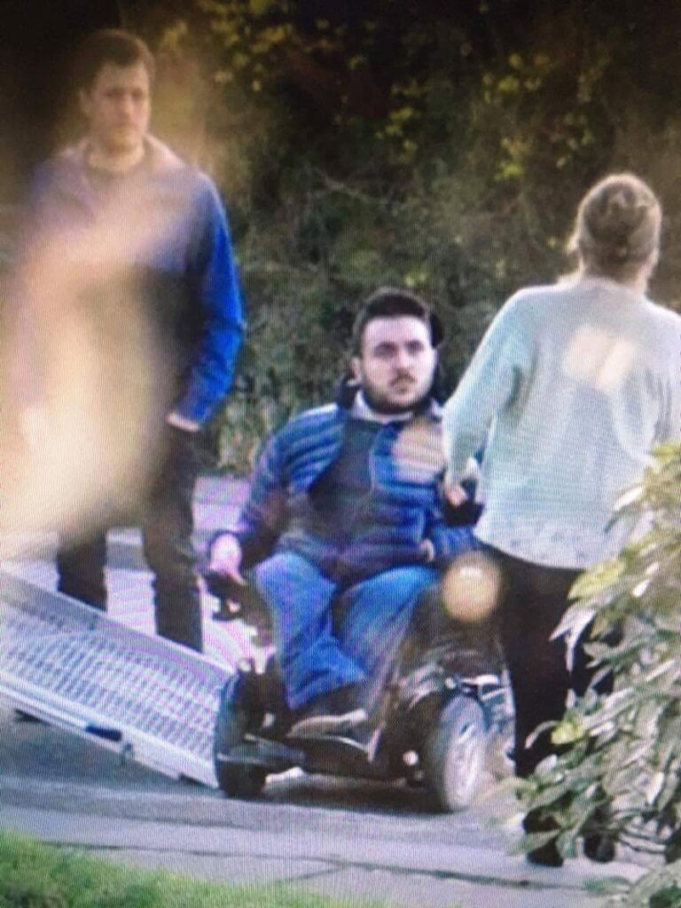En mand i en elektrisk kørestol bruger en bærbar rampe på en sti, ledsaget af en kvinde, der sidder vendt mod ham, og en anden mand, der står i nærheden. I baggrunden ses træer og grønne områder.